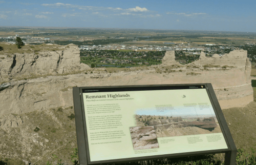 A scenic view of Remnant Highlands with informational sign, overlooking a valley and distant town under a blue sky.