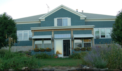 A blue house with a porch, surrounded by greenery and flowers, under a clear sky.