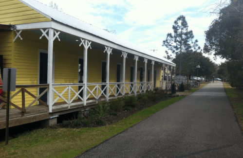 A yellow building with a porch alongside a paved path, surrounded by greenery and trees.