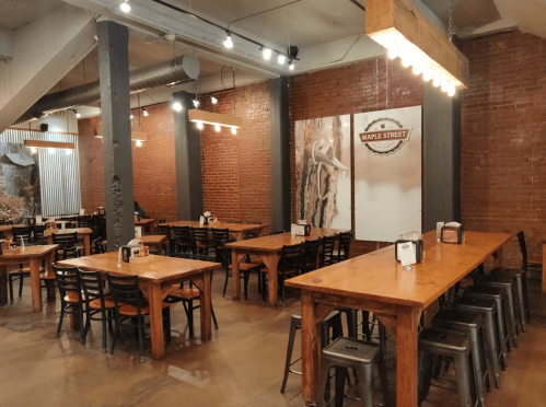Interior of a restaurant with wooden tables, metal chairs, and brick walls, featuring a rustic decor style.