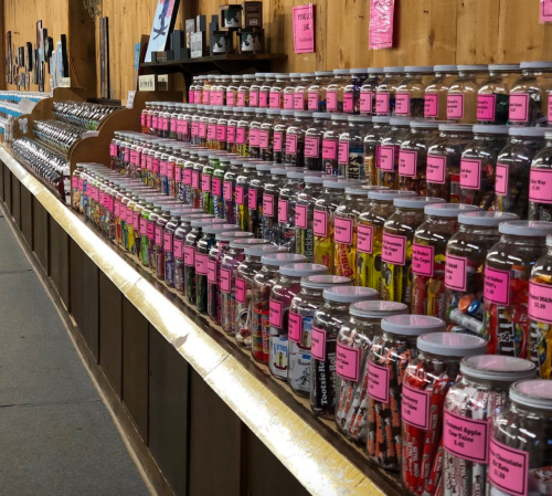 A colorful display of jars filled with various candies, each labeled with bright pink tags, arranged on wooden shelves.