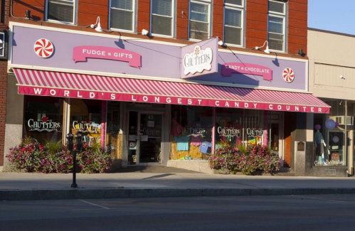 A colorful candy store with a striped awning, featuring "World's Longest Candy Counter" sign and vibrant flower displays.