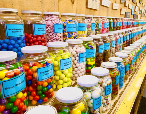 A colorful display of jars filled with various types of candies, labeled with names, arranged on a wooden shelf.