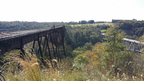 An old, rusted bridge spans a valley, surrounded by lush greenery and distant hills under a clear sky.