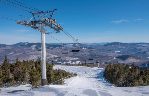 A ski lift towers over a snowy mountain landscape, with skiers on the slope and distant mountains under a clear blue sky.