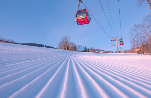 A ski resort scene featuring a cable car above freshly groomed snow and trees under a clear blue sky.