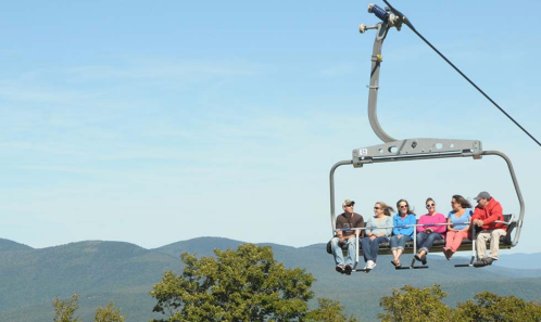 A group of people on a ski lift, surrounded by mountains and a clear blue sky.