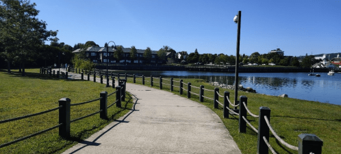 A paved path along a waterfront, lined with wooden posts and greenery, leading to buildings in the distance under a clear blue sky.