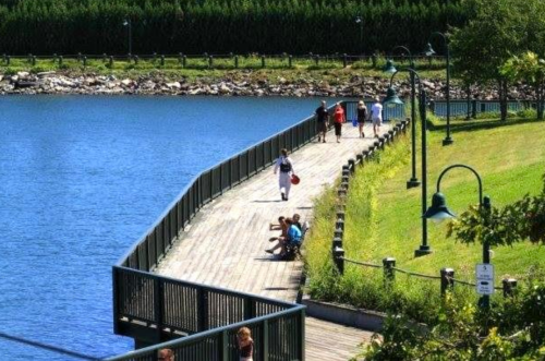 A scenic boardwalk by a lake with people walking and sitting, surrounded by greenery and lampposts.
