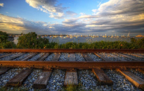 A scenic view of a railway track leading to a harbor filled with sailboats under a cloudy sky.