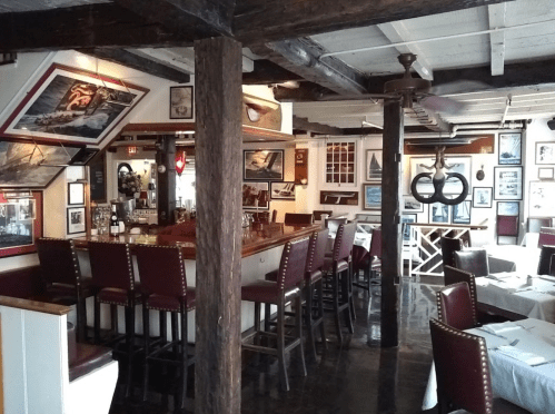 Cozy restaurant interior featuring a bar, wooden beams, and framed nautical-themed artwork on the walls.