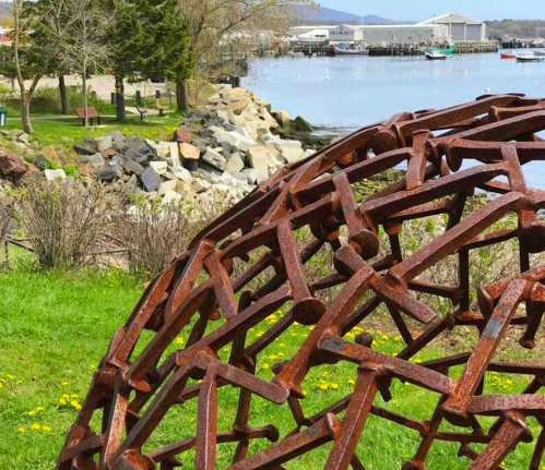 A rusted metal sculpture in a grassy area, overlooking a calm waterfront with boats and rocky shoreline in the background.