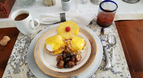 A beautifully arranged breakfast plate with eggs Benedict, roasted potatoes, a pineapple slice, coffee, and juice.