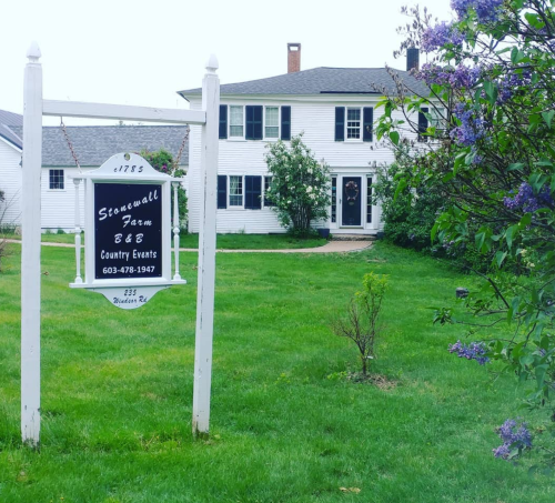 A white two-story house with a sign reading "Stonewall Farm B&B" in a green yard with blooming flowers.