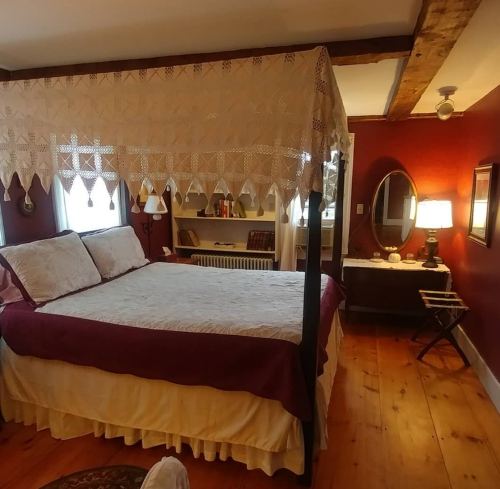 Cozy bedroom with a four-poster bed, warm red walls, wooden floors, and vintage decor. Soft lighting creates a welcoming atmosphere.