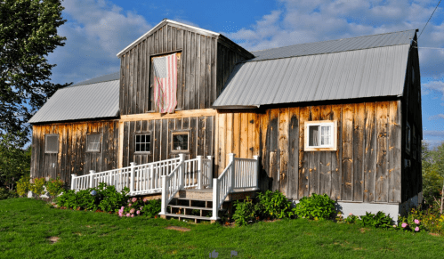 A rustic wooden barn with a metal roof, featuring an American flag and surrounded by green grass and flowers.