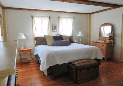 Cozy bedroom with a large bed, decorative pillows, wooden furniture, and soft natural light from window curtains.