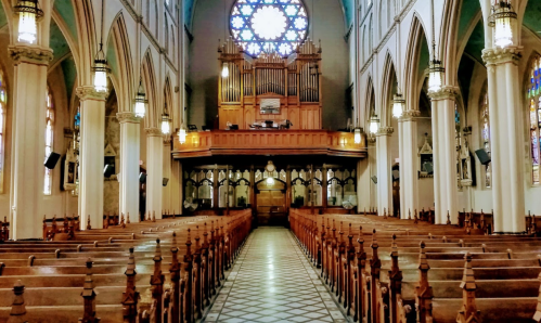 Interior of a grand church featuring wooden pews, a large organ, and a colorful stained glass window.