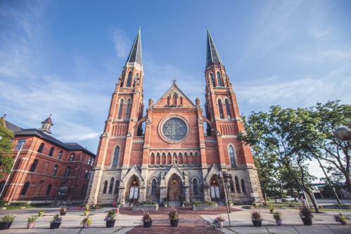 A grand brick church with two tall spires and a large rose window, surrounded by greenery and flowers.
