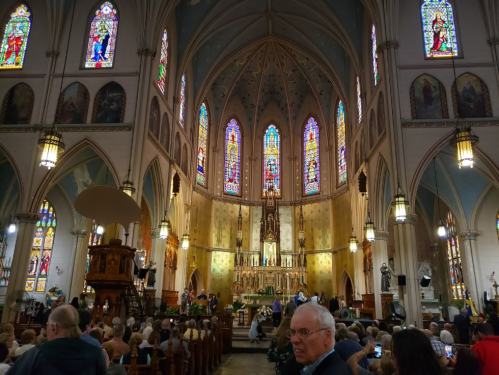 Interior of a grand church with stained glass windows, a congregation, and a central altar, creating a serene atmosphere.