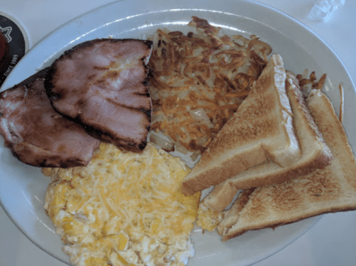 A plate of breakfast featuring ham, scrambled eggs with cheese, hash browns, and toasted bread.