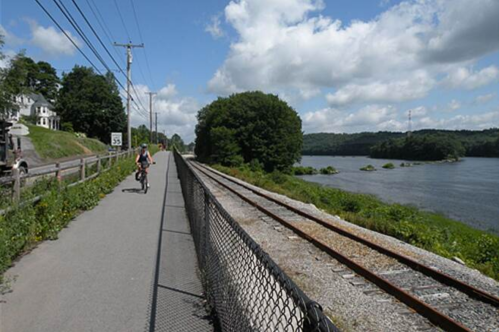 A cyclist rides along a path beside a river, with trees and a railway track in the background under a blue sky.