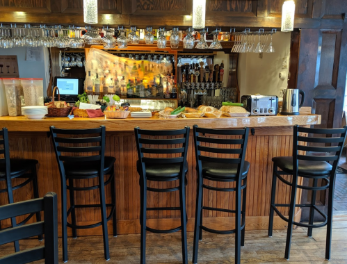 A wooden bar with high stools, drinks on tap, and a variety of food items displayed on the counter.