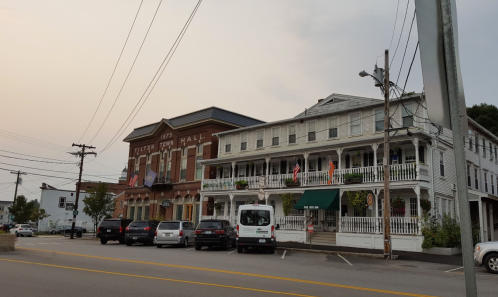Historic town hall building next to a multi-story white building, with parked cars and flags displayed.