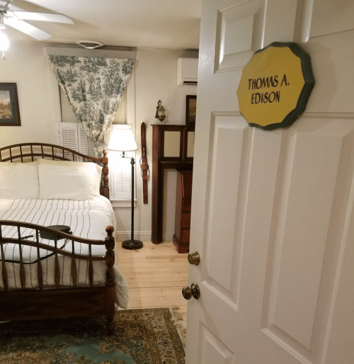 A cozy bedroom with a bed, lamp, and decor, featuring a door sign that reads "Thomas A. Edison."