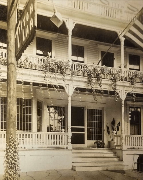 Historic photo of a charming inn with a sign, porch, and decorative plants.
