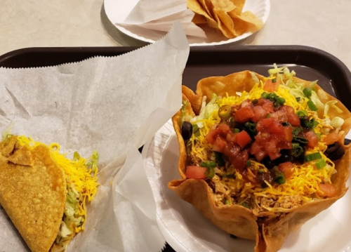 A tray with a taco in paper and a taco salad bowl topped with lettuce, cheese, tomatoes, and olives.