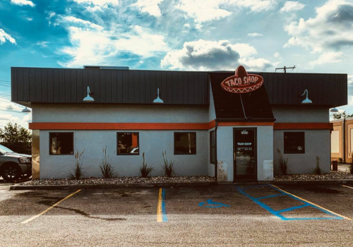 A taco shop with a colorful sign, gray exterior, and parking lot under a partly cloudy sky.