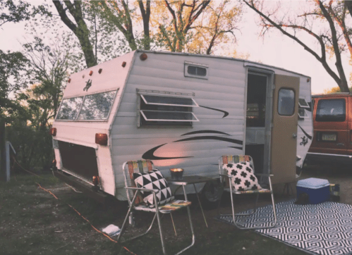 A vintage camper parked in a campsite, surrounded by trees, with two chairs and a patterned rug outside.