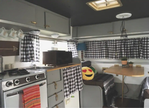 Cozy camper interior with gray cabinets, checkered curtains, a kitchenette, and a cheerful emoji pillow on a sofa.