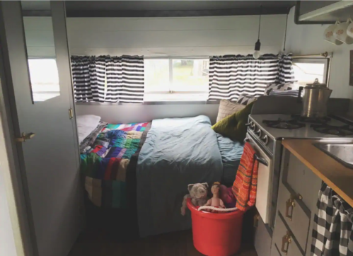 Cozy bedroom in a camper with a bed, colorful quilt, and a small kitchen area, featuring striped curtains and a pet dog.