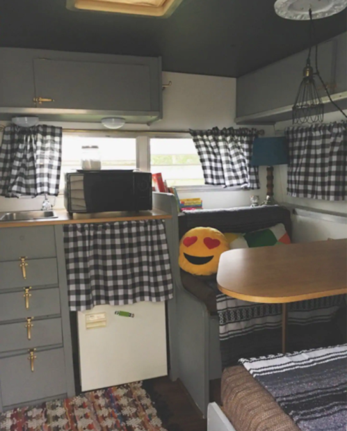 Cozy camper interior with checkered curtains, a kitchenette, a dining table, and a smiling emoji pillow on the couch.