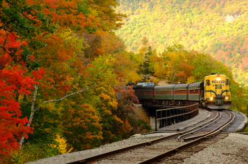 A vintage train travels along a curved track surrounded by vibrant autumn foliage in a scenic landscape.