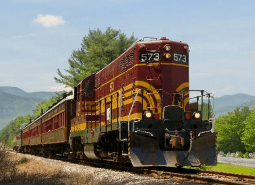 A vintage red and yellow train, number 573, travels along a scenic track with mountains in the background.