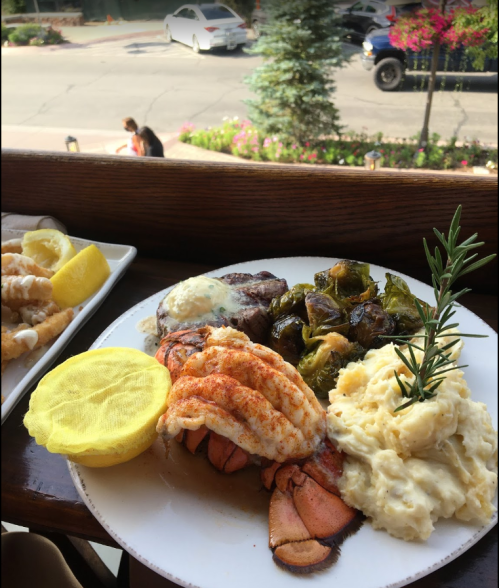 A plate of seafood and steak with mashed potatoes, Brussels sprouts, and lemon, set against a scenic outdoor backdrop.