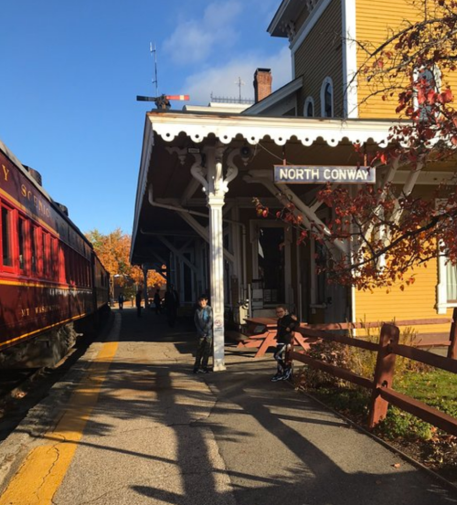 A train station with a sign reading "North Conway," featuring two children on the platform and autumn foliage nearby.