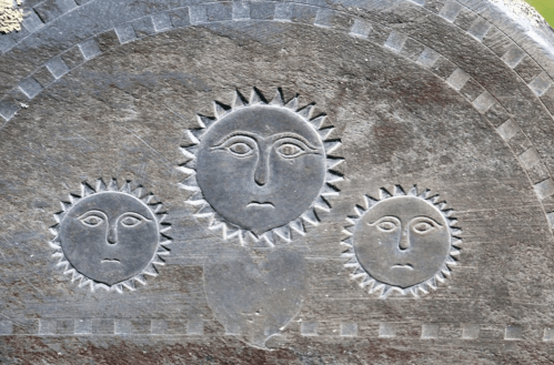 Carved stone featuring three sun-like faces with distinct expressions, set against a textured background.