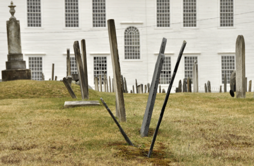 Gravestones at a cemetery with a historic building in the background, set against a grassy landscape.