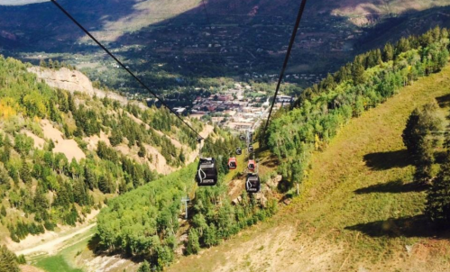 Aerial view of a gondola ride over a lush green valley and a small town below, surrounded by mountains.