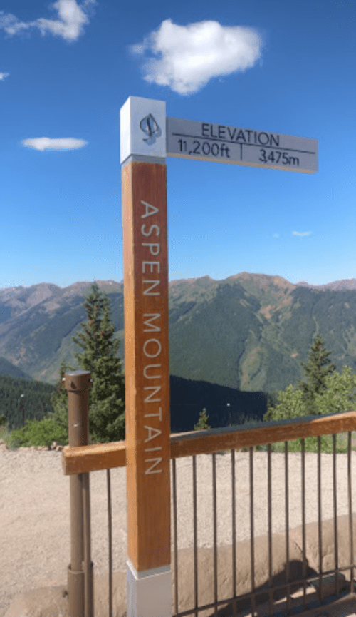 Signpost indicating the elevation of Aspen Mountain at 11,200 feet (3,475 meters) with a scenic mountain backdrop.