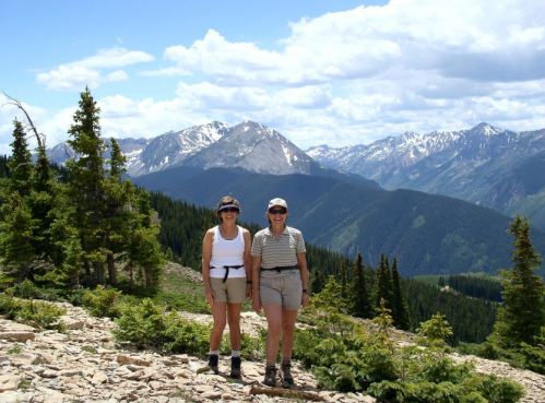 Two women stand on a rocky trail with mountains and trees in the background, enjoying a sunny day outdoors.