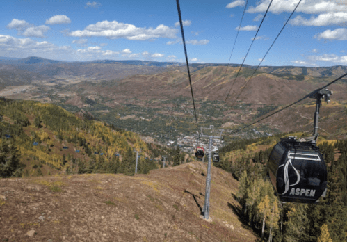 A scenic view from a gondola in Aspen, showcasing mountains, valleys, and a clear blue sky with scattered clouds.
