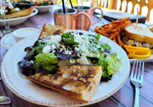 A grilled flatbread topped with mixed greens and feta cheese, served with a side of sweet potato fries.