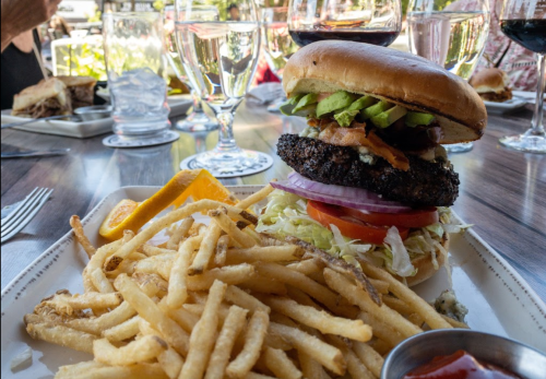 A close-up of a burger with avocado, bacon, and toppings, served with a side of golden fries and a slice of orange.