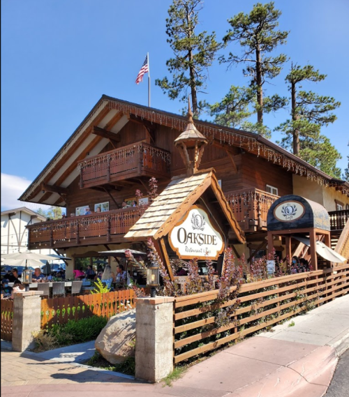 A charming wooden restaurant with balconies, surrounded by trees and a clear blue sky. Outdoor seating is visible.