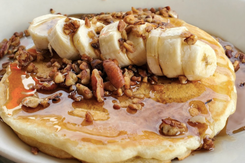 A fluffy pancake topped with sliced bananas, chopped nuts, and drizzled with syrup.